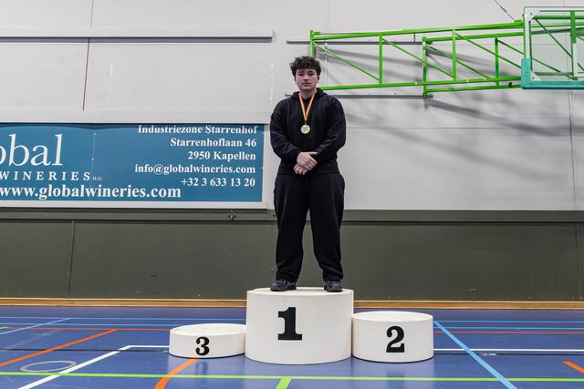 Jonge mannelijke sporter staat trots op het winnaarspodium met een gouden medaille in een sporthal.