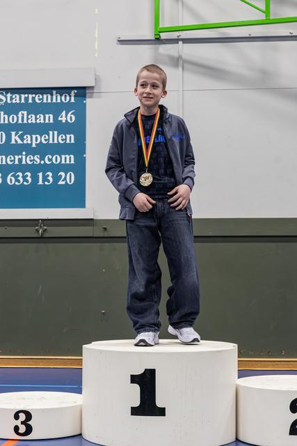 Lachende jongen staat trots op het eerste podium met een gouden medaille aan een driekleurig lint in een sporthal.