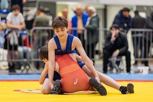 Jongen in blauw tenue houdt tegenstander in rood onder controle op de gele mat, met geconcentreerde blik.