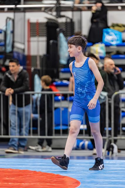 Jonge jongen in blauw wrestlingpak loopt zelfverzekerd over de mat tijdens een worstelwedstrijd, toeschouwers op de achtergrond.