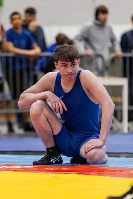 Jonge worstelaar in blauw trikot hurkt op de mat en kijkt gefocust en vastberaden in de camera.