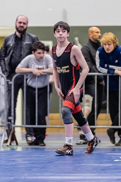 Jonge worstelaar in zwart-rood tenue staat waakzaam op de blauwe mat, geconcentreerde blik, coaches kijken toe achter de hekken.