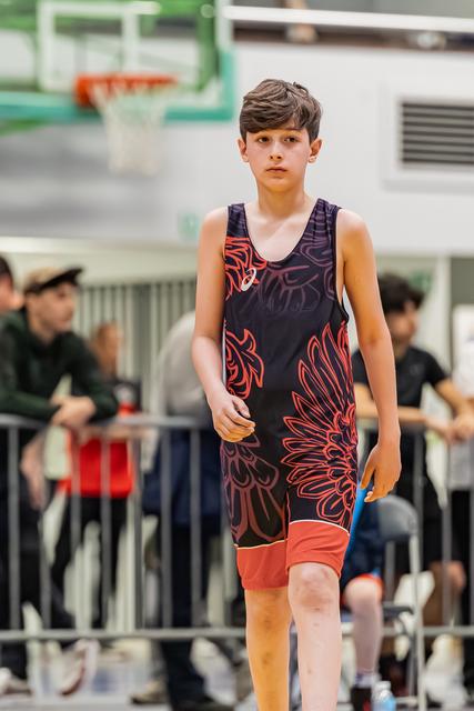 Jonge jongen in ASICS singlet loopt gefocust over de gymzaalvloer tijdens een worstelevenement.