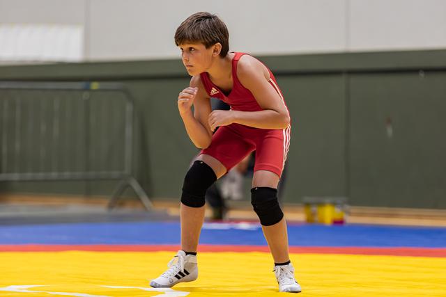 Jonge worstelaar in rood Adidas tenue staat geconcentreerd en alert in vechthouding op de wedstrijdmat.