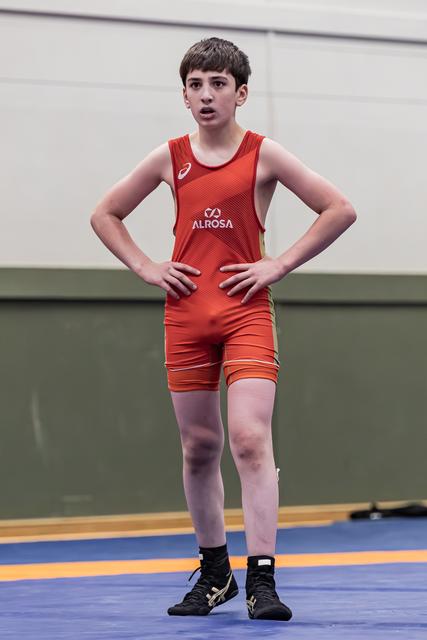 Jonge worstelaar in rood Alrosa tenue staat met handen op de heupen, waakzame blik, op blauwe mat.