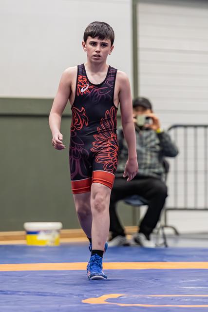 Jonge jongen in zwart-rood singlet loopt met een geconcentreerde, vastberaden blik over de mat tijdens een worstelevenement.