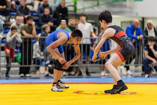 Twee jongens in worstelhouding staan geconcentreerd tegenover elkaar op een gele mat, met publiek op de achtergrond.