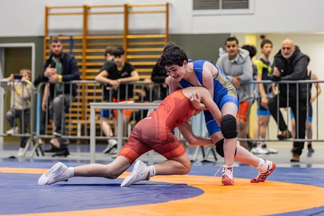 Twee jonge worstelaars strijden op de mat; de blauwe atleet dringt agressief vooruit terwijl de rode tegenstander laag stand houdt.