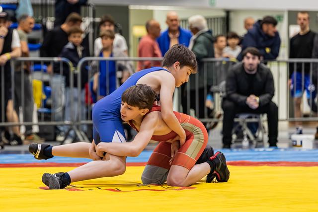 Twee jongens in blauwe en rode wrestlingpakken verwikkeld in een fel gevecht op een gele mat, met toeschouwers op de achtergrond.