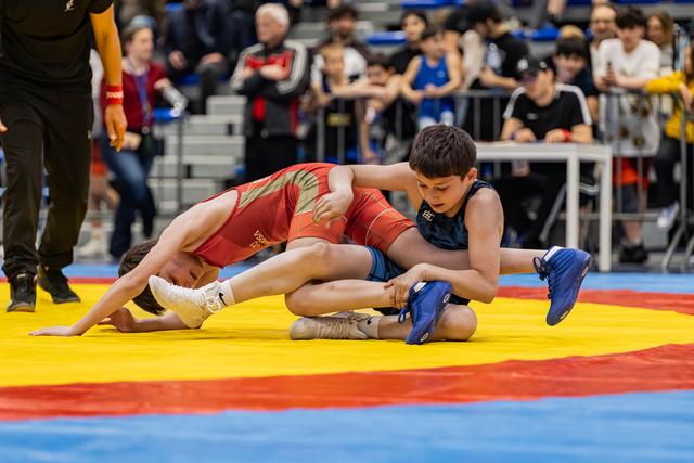Twee jonge worstelaars strijden op een kleurrijke mat, terwijl de blauwe atleet zijn tegenstander in het rood bij het been grijpt.
