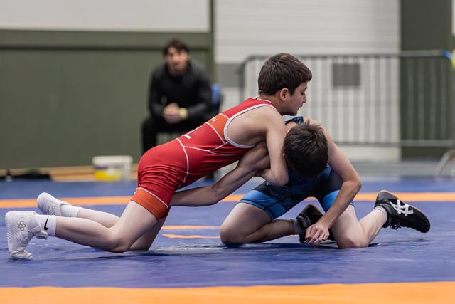 Twee jonge worstelaars strijden fel op een blauwe mat, de een in het rood heeft de overhand terwijl een coach toekijkt.