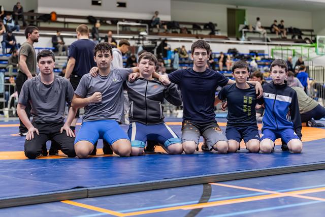 Zes jonge mannelijke worstelaars knielen samen op een blauwe mat, armen om elkaar heen, en poseren vol zelfvertrouwen voor een teamfoto.