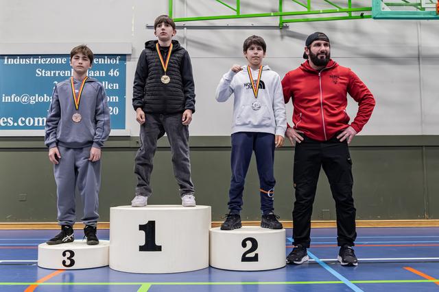 Drie jonge worstelaars staan met medailles op het podium; een volwassen coach in het rood staat trots naast hen.