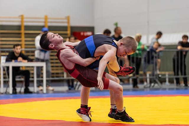Jonge worstelaar in rood tenue tilt tegenstander op van de mat tijdens een intense wedstrijd in een sporthal.