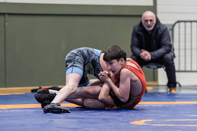 Jonge worstelaar in rode singlet houdt een verdedigende positie op de mat terwijl tegenstander in blauw druk uitoefent, coach kijkt toe.