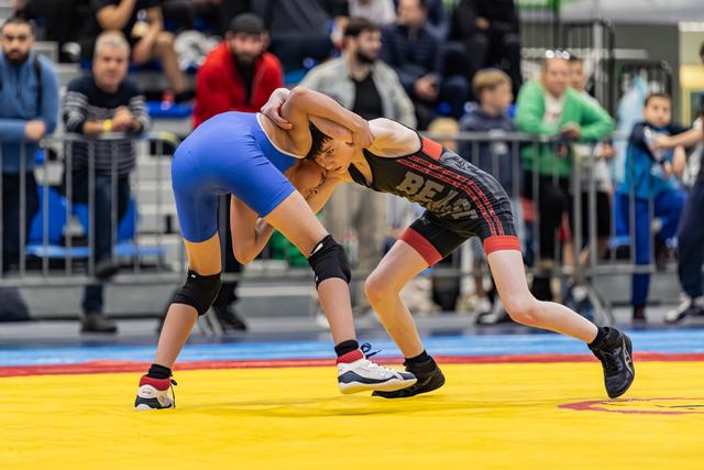 Twee jonge worstelaars grijpen elkaar stevig vast in een intense clinch, één in blauw en één in een zwart Bears-tenue, op een gele mat.