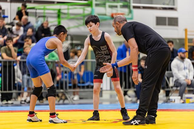 Twee jonge worstelaars schudden elkaar de hand voor een wedstrijd, begeleid door een scheidsrechter op de gele mat in een drukke sporthal.