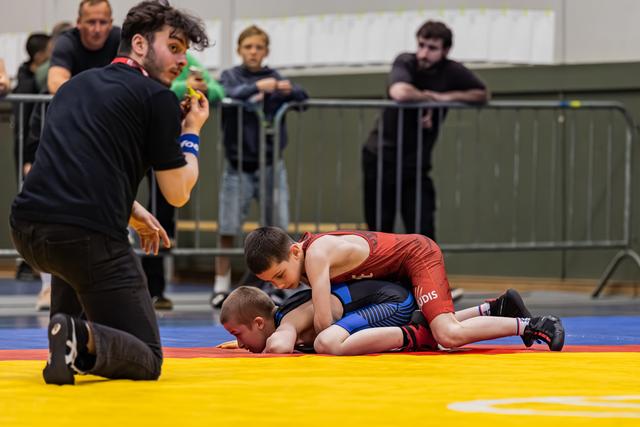 Jonge worstelaar in rood tenue pinnt tegenstander op de mat terwijl de scheidsrechter aandachtig toekijkt in een sporthal.