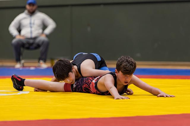 Twee jonge jongens worstelen op een gele mat, één houdt de ander neer terwijl een coach aandachtig toekijkt.