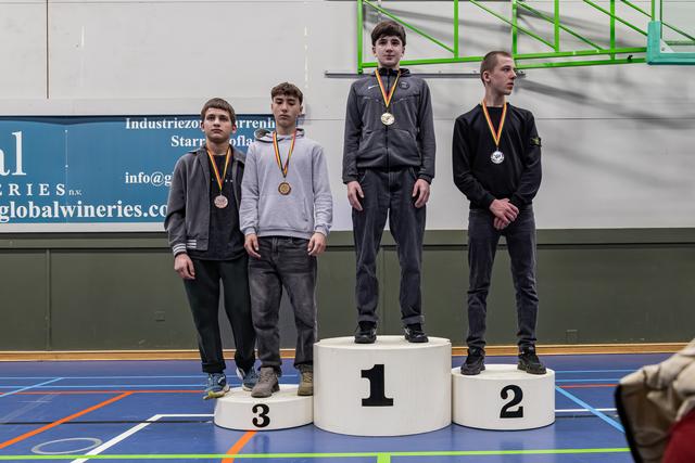 Vier jonge mannelijke worstelaars staan op een podium met medailles, de gouden medaillewinnaar staat het hoogst op positie 1.