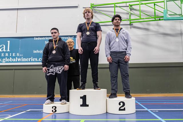 Drie tieners staan op het podium met medailles, begeleid door een coach in een sporthal.