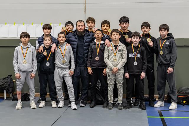 Jonge worstelaars tonen trots hun gouden medailles samen met hun coach in een sporthal na een succesvol toernooi.