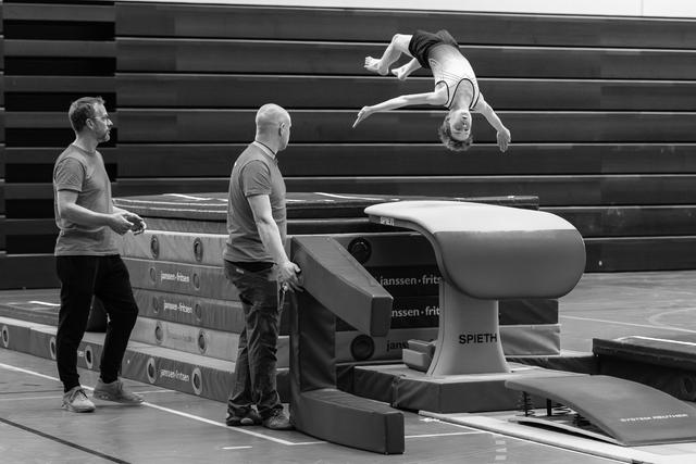 Athlete performs inverted backflip over vaulting table while two coaches stand ready with safety mats during training