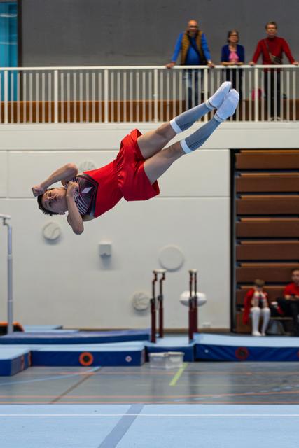 Athlete in red uniform performs a backwards somersault in mid-air above the floor exercise mat while spectators watch from balcony