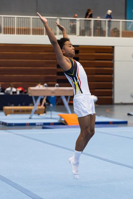 Athlete performing floor routine with arms raised during jump, white leotard with colorful design, indoor gymnasium