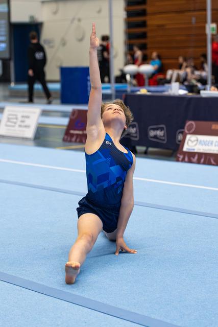 Young gymnast performs an expressive floor routine in kneeling position with one arm raised, looking upward on blue mat