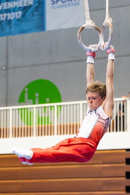 Young athlete demonstrating an L-sit position on still rings during indoor training, wearing red and white uniform