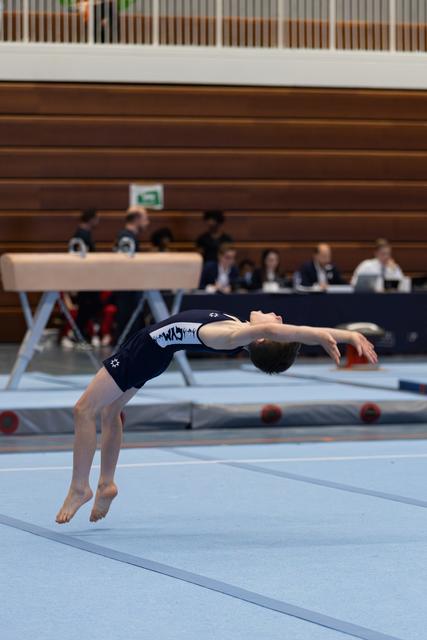 Young athlete performs dramatic backbend during floor routine, arching backwards mid-air above competition mat