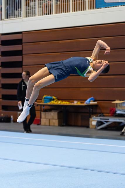 Young athlete performing a dramatic backward arch during floor routine, white socks visible, wooden bleachers in background
