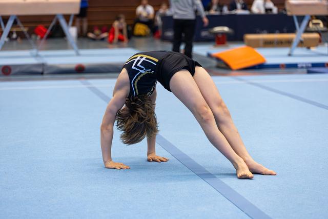 Young athlete performs a bridge backbend on the floor mat, demonstrating flexibility during a routine at an indoor event.