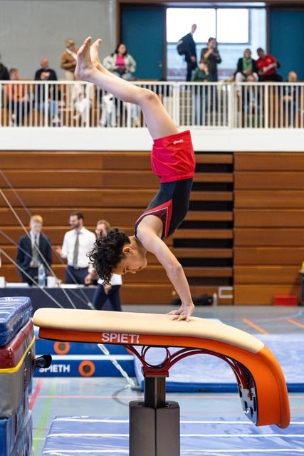 Female gymnast executes a handstand on vault apparatus during competition, displaying strength and balance with judges watching