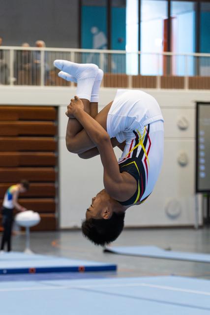 Athlete mid-backflip in tucked position, demonstrating impressive flexibility and control during floor routine in sports facility