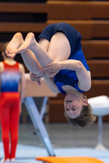 Young athlete performs a tucked backflip mid-air during routine, displaying intense focus and athleticism in the arena