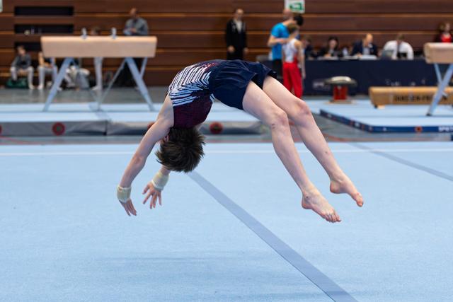 Athlete performs a backflip mid-air during floor routine, demonstrating flexibility and control in an indoor arena