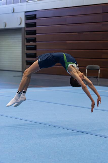 Athlete performing a backward arch flip mid-air on floor exercise mat in training facility