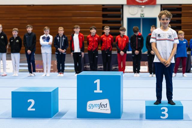 Bronze medalist stands proudly on third place podium wearing white tracksuit while other competitors line up in background