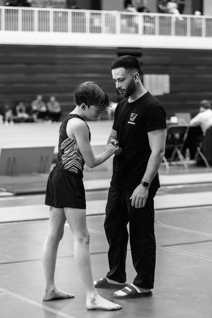 Coach in black attire comforting young athlete in sparkly leotard on gymnasium floor, displaying supportive mentorship