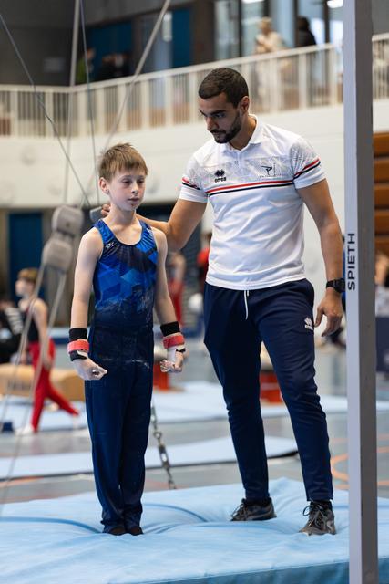 Coach in white polo shirt walking beside young athlete in blue leotard on competition floor, sharing an encouraging moment