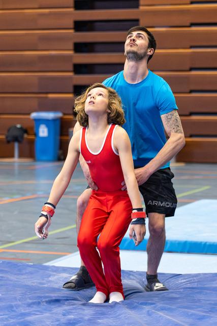 Coach guiding young athlete in red singlet, both looking upward during training session in indoor sports hall