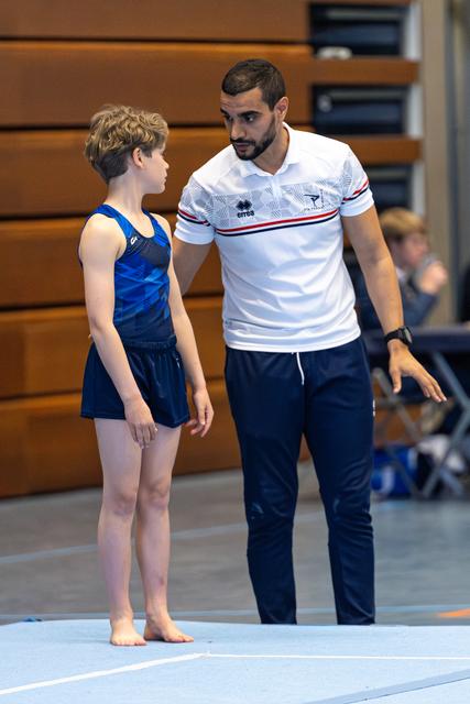 Coach in white polo speaks with young athlete in blue leotard on floor mat during training session