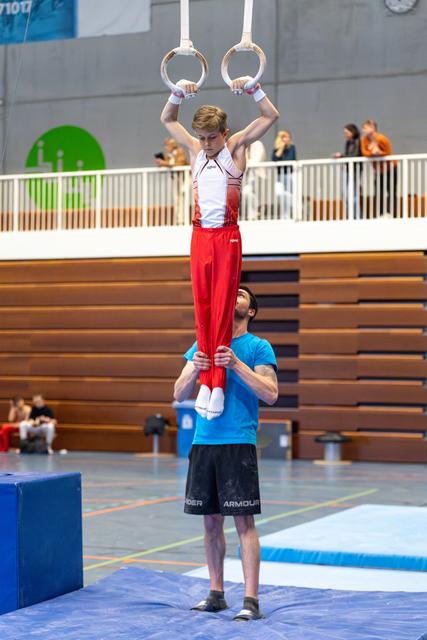 Coach spotting a young athlete performing a strength hold on rings during training at an indoor sports hall