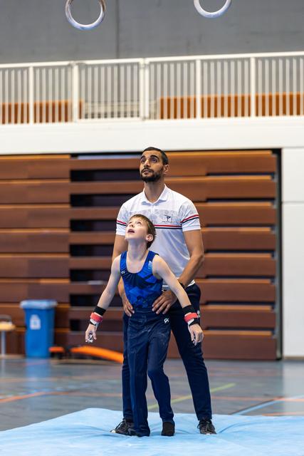 Coach steadies young athlete in blue leotard as both gaze upward with intense focus during training session in gymnasium