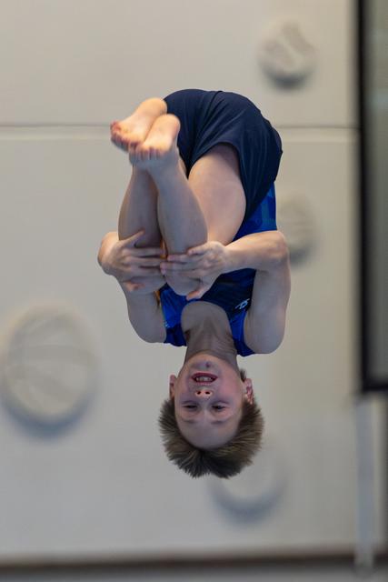 Athlete performing an inverted tuck during a dive, captured mid-flight against a neutral indoor backdrop