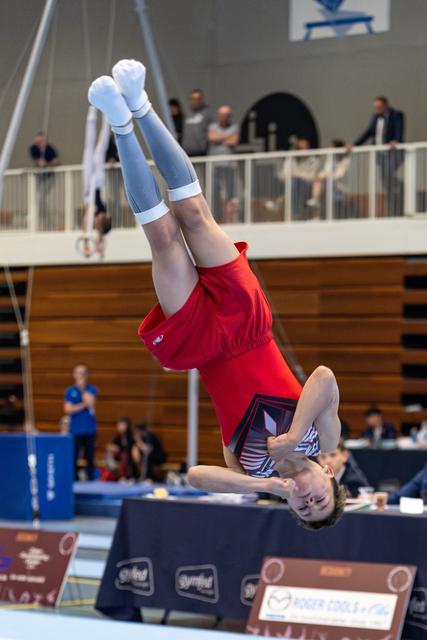Female gymnast performs inverted handstand on floor exercise mat during competition, legs extended vertically in red leotard