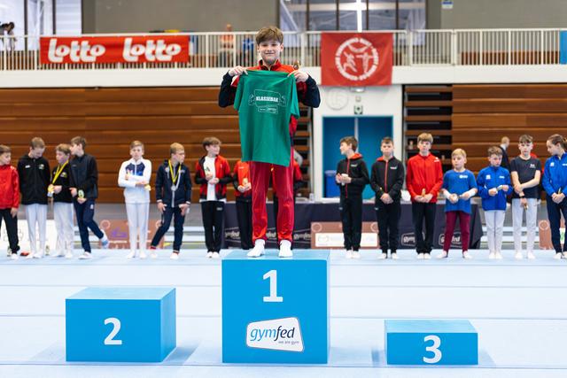 Winner in green uniform stands on first place podium with raised fists while other competitors stand in background