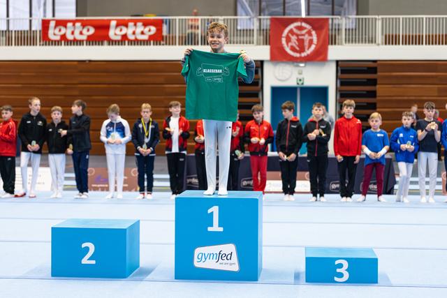 Young athlete on first place podium holding up a green team shirt while fellow competitors with medals stand behind